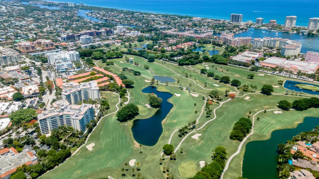 Aerial view over Mizner Park in Boca Raton, looking down the palm-lined central green to the bandshell, framed by coral-pink Mediterranean-style buildings.
