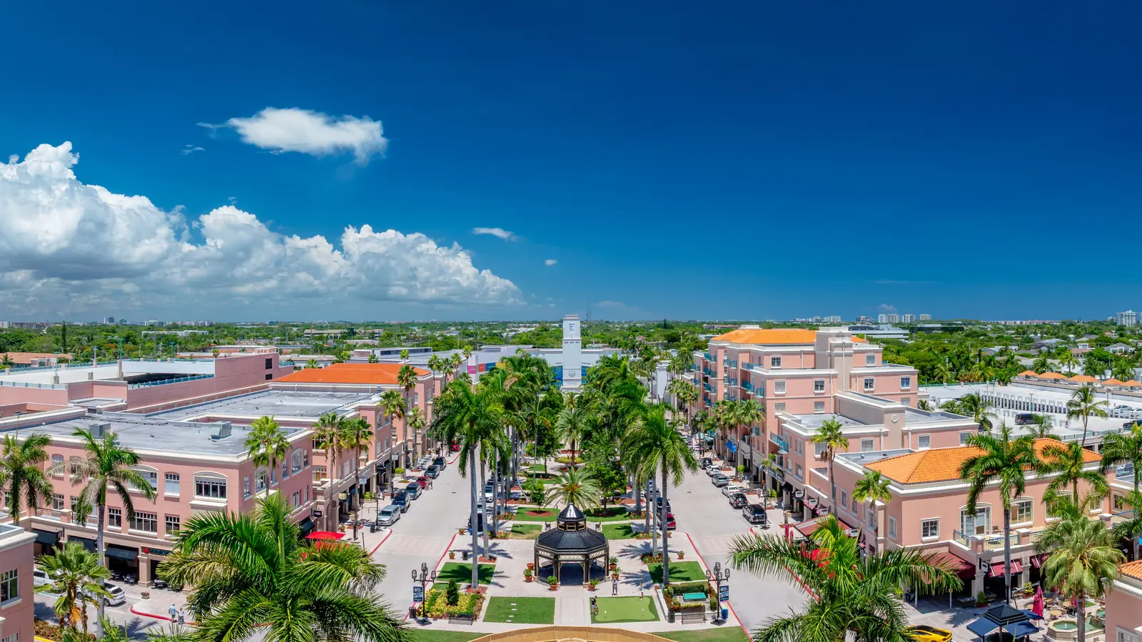 Aerial view over Mizner Park in Boca Raton, looking down the palm-lined central green to the bandshell, framed by coral-pink Mediterranean-style buildings.
