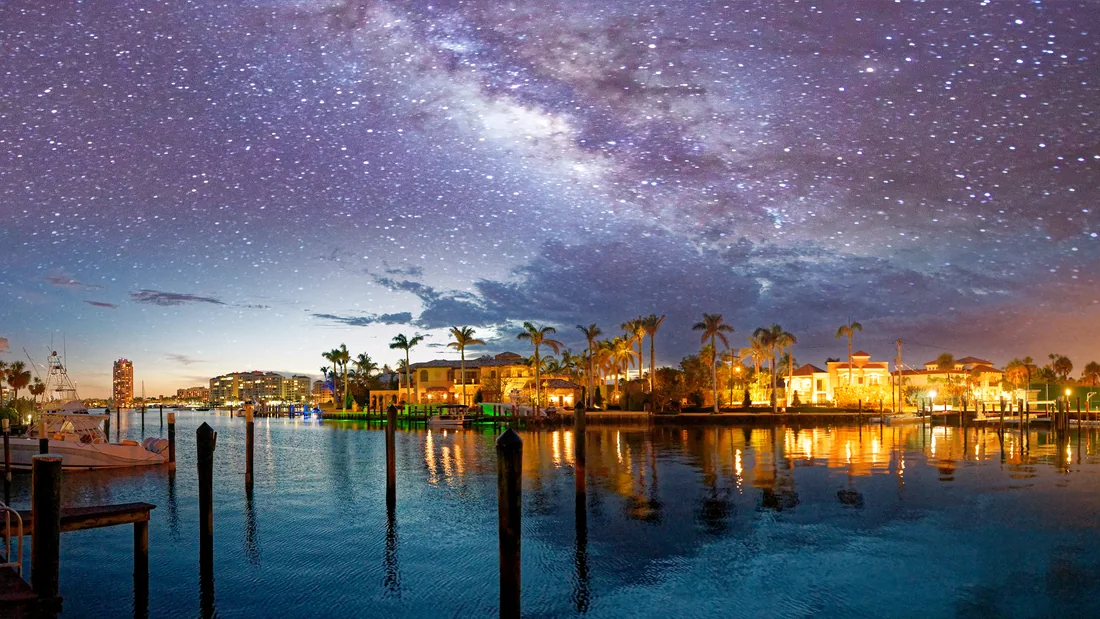 A South Florida marina at twilight under a star-filled sky, with wooden dock pilings and moored boats in the foreground and calm water reflecting the lights of low-rise waterfront buildings and palm trees along the far shore.