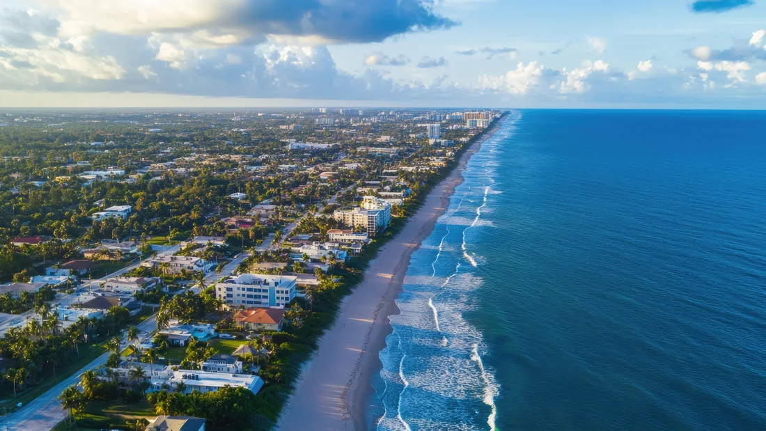 Aerial view of a South Florida coastal strip, with the Intracoastal Waterway on one side, single-family neighbourhoods and oceanfront condominium towers in the middle, and the Atlantic Ocean on the other.