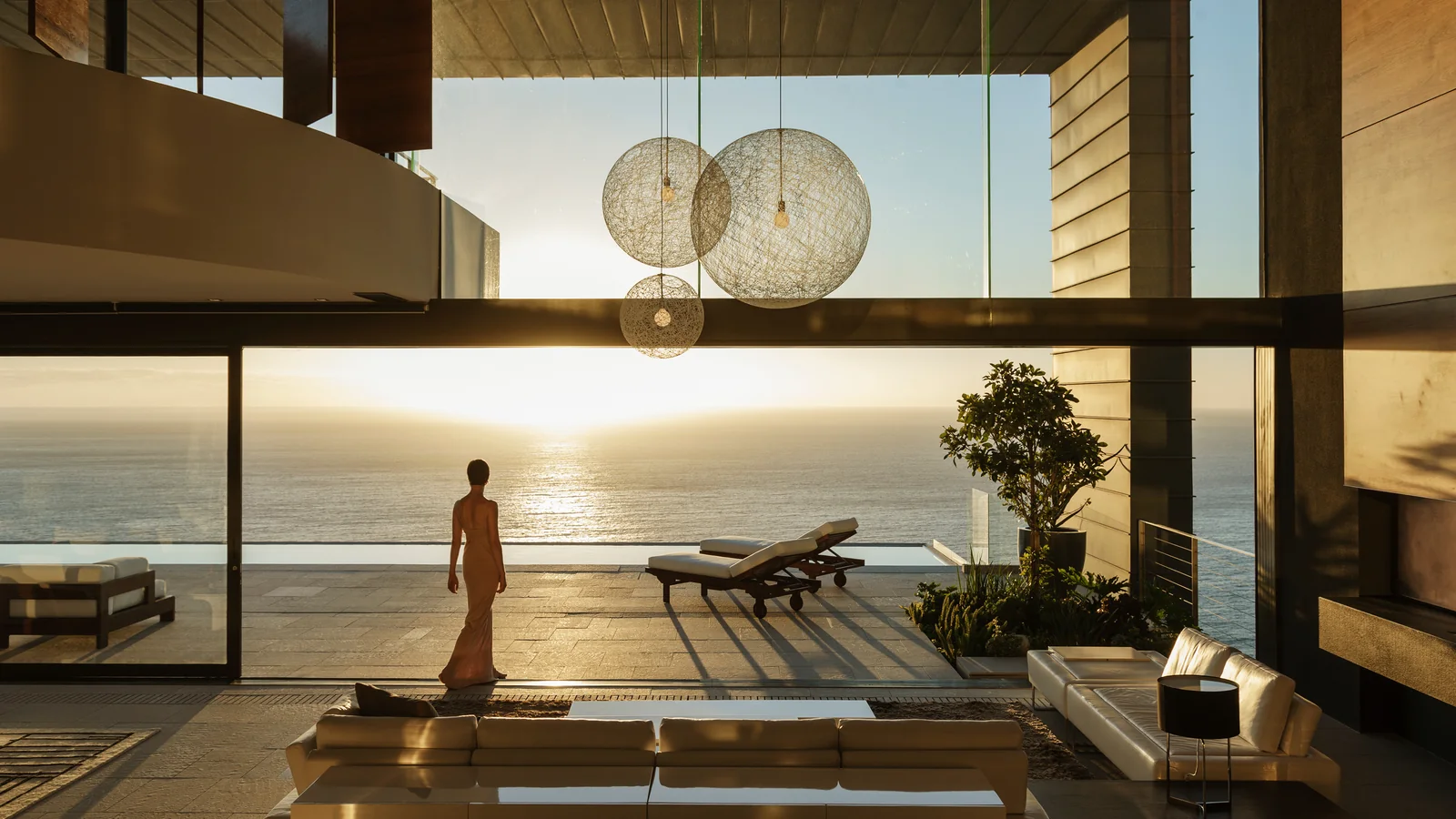 Interior of a luxury home at sunset, looking through floor-to-ceiling glass onto an ocean-facing terrace, with globe pendant lights overhead.
