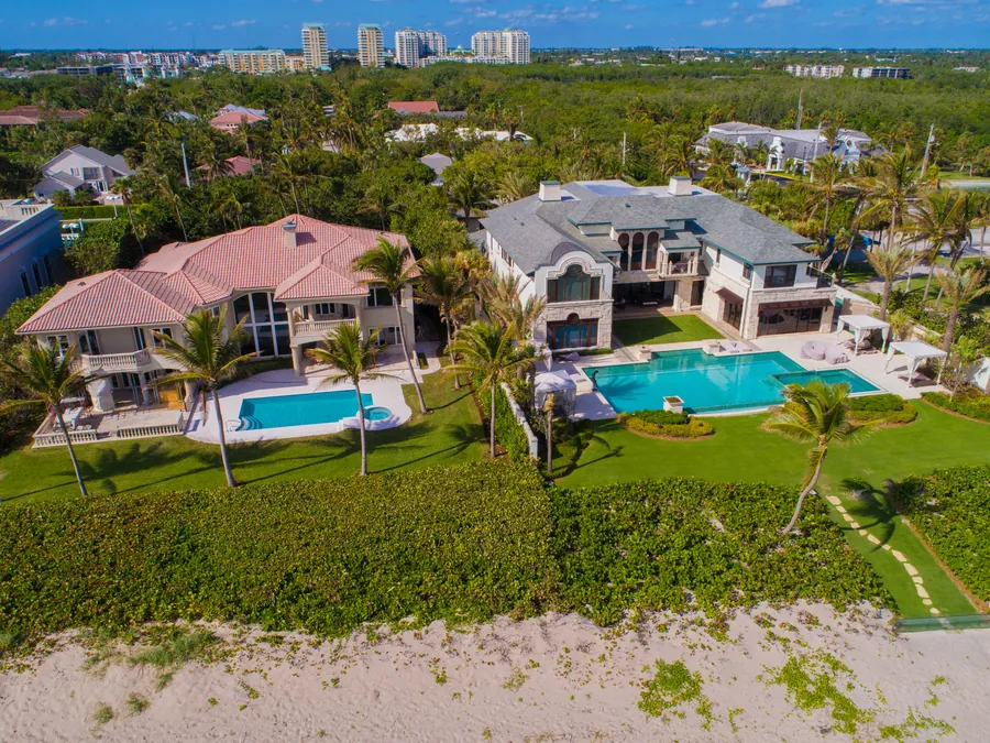 Aerial view of a row of oceanfront estates on a South Florida beach, with swimming pools and manicured lawns visible from above between the houses and the sand.
