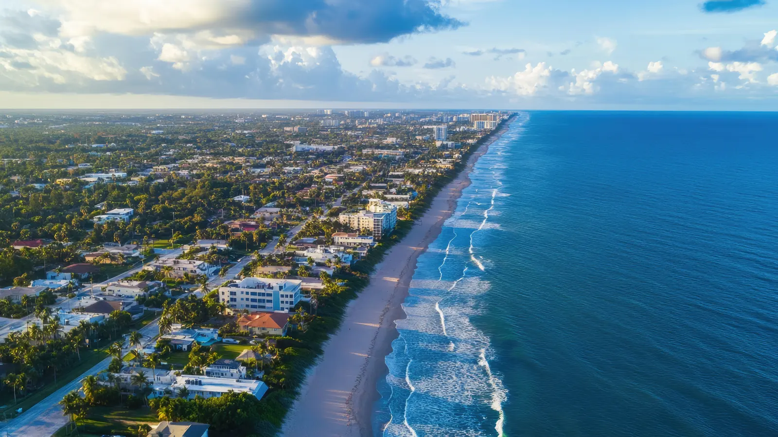 Golden-hour aerial view along a South Florida barrier island: waves breaking along a wide sand beach, low-rise homes and small condominium buildings on palm-lined streets, and the coastline receding toward a distant skyline.