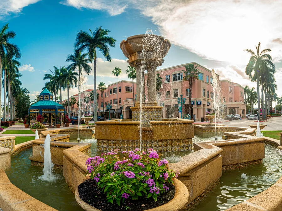 Mizner Park in Boca Raton at street level, showing the central fountain, coral-pink Mediterranean buildings and rows of royal palms with people on the walkways.
