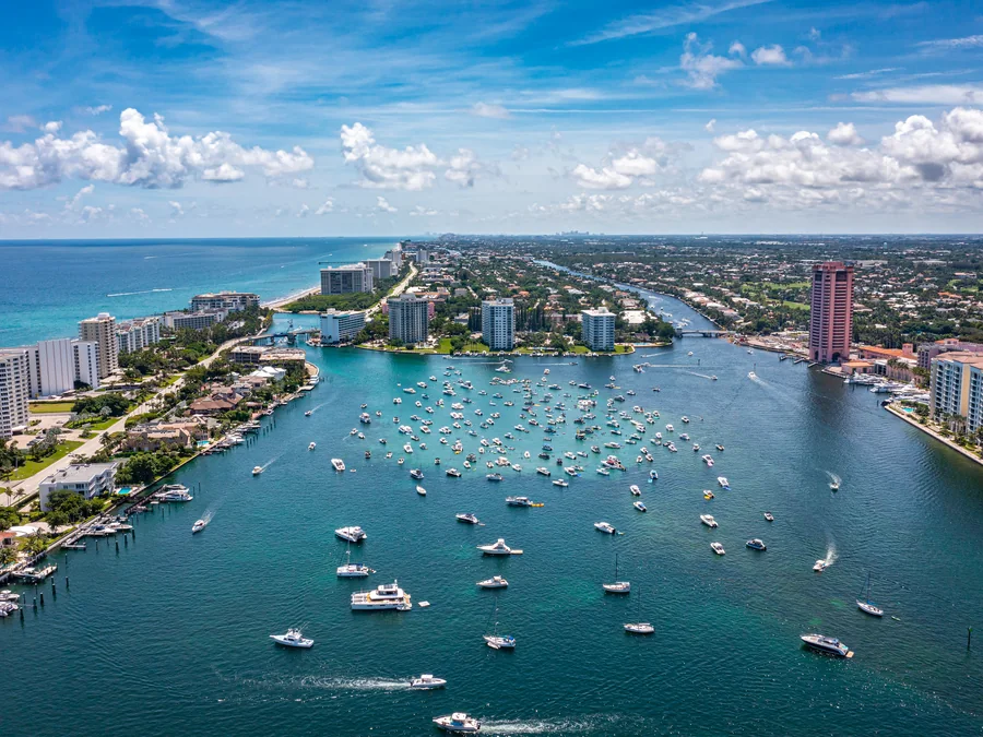 Aerial view of Lake Boca Raton and the Boca Raton Inlet, with dozens of boats anchored in turquoise water, condominium towers along both shores and the Atlantic Ocean beyond.