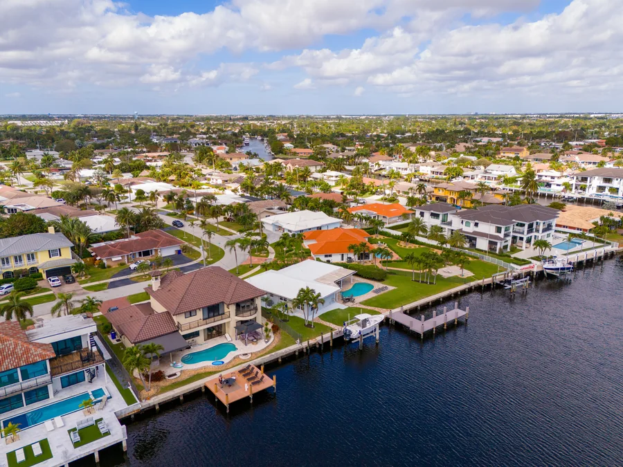 Aerial view over a South Florida Intracoastal waterway: single-family homes with swimming pools, private docks and boat lifts line the near bank, with tile-roofed residential streets and palm trees stretching inland.
