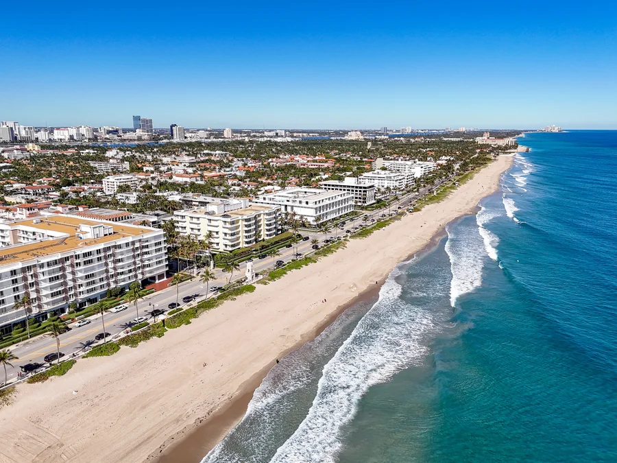 Aerial view of a South Florida oceanfront at midday: waves breaking along a wide sand beach, a palm-lined coastal road and low-rise white residential buildings with terracotta roofs.