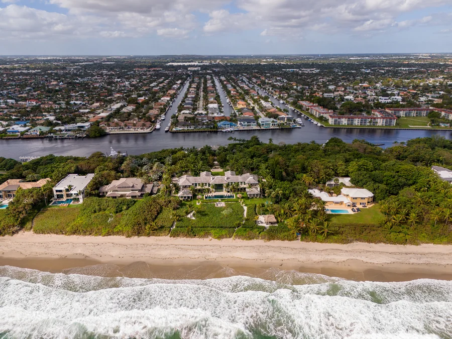 Aerial view of a South Florida barrier island: Atlantic surf breaking on the beach in the foreground, a row of oceanfront estates with swimming pools behind the dune, the Intracoastal Waterway beyond them, and canal-front streets with private docks in the distance.